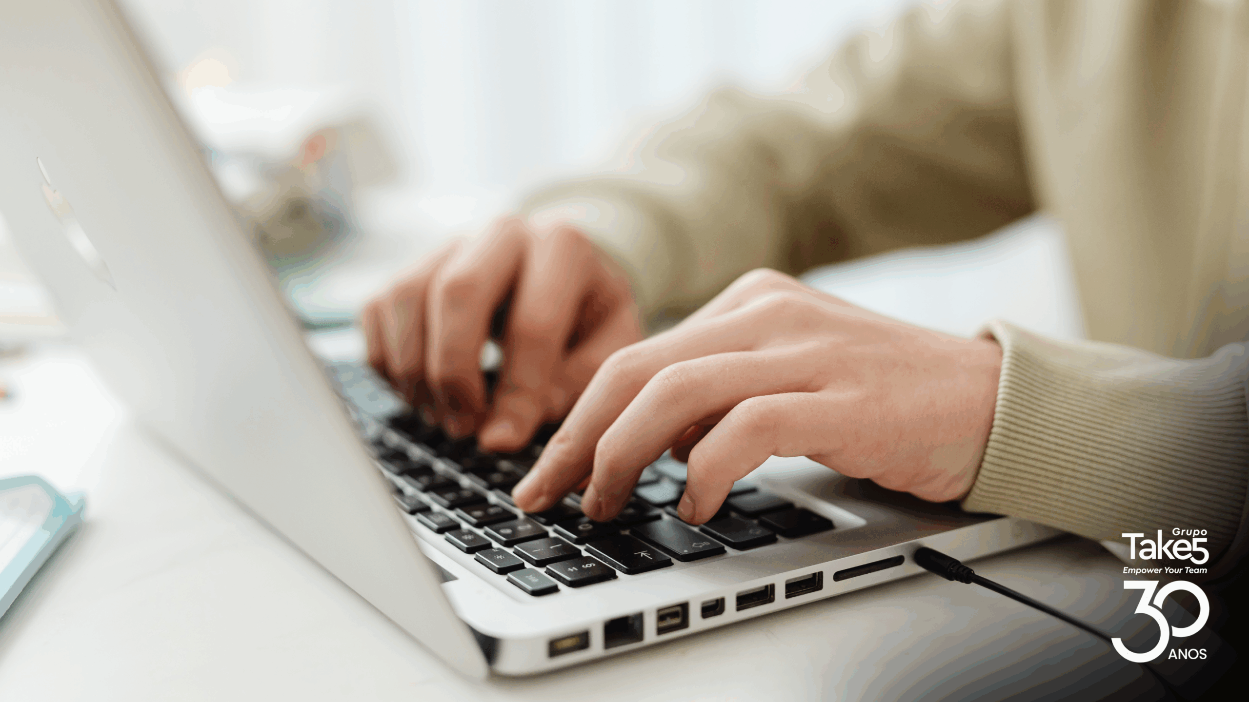 Mãos digitando em notebook em ambiente de trabalho, representando uso de tecnologia para aprendizagem digital e educação corporativa, com identidade do Grupo Take 5.
