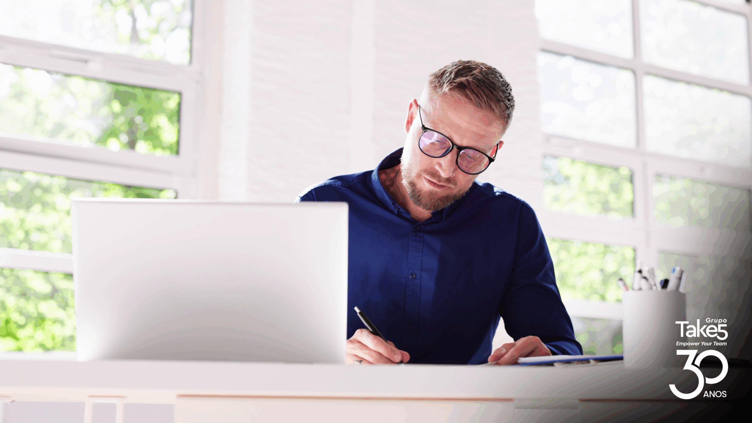 Profissional trabalha em notebook e faz anotações à mesa em ambiente corporativo iluminado, representando aprendizagem contínua e educação corporativa do Grupo Take 5.